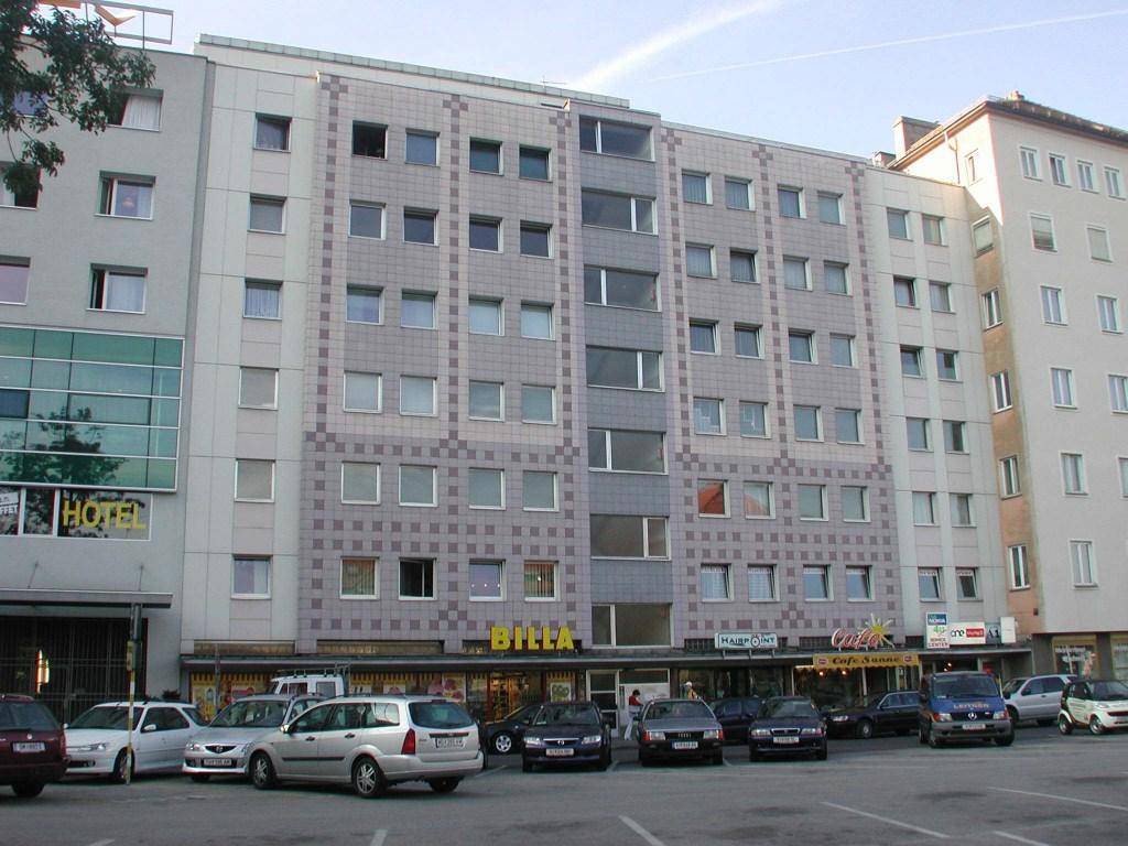 Mehrstöckiges Gebäude mit Geschäften im Erdgeschoss und geparkten Autos davor, zeigt städtische Architektur.