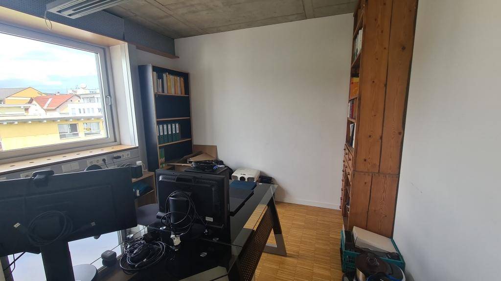 Einzelbüro mit einem Schreibtisch, Computermonitoren und Holzregalen, das durch ein großes Fenster hell beleuchtet wird.