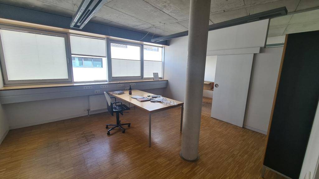 Leerer Büroraum mit Holzparkettboden, einem einzelnen Schreibtisch und großen Fenstern, die viel Licht hereinlassen.