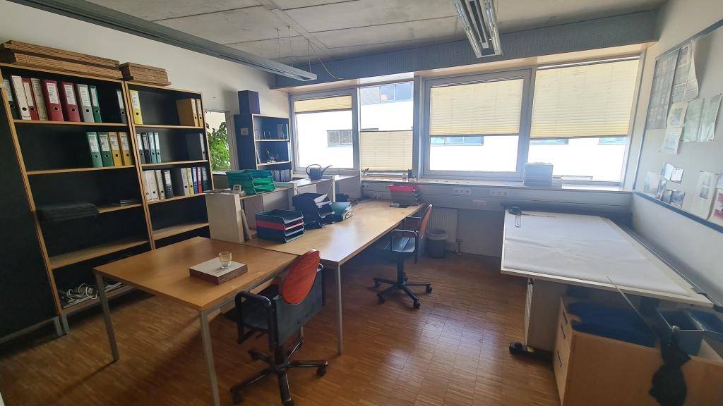 Helles Büro mit mehreren Arbeitsplätzen, Holzparkettboden und großen Fenstern, die viel Tageslicht hereinlassen.