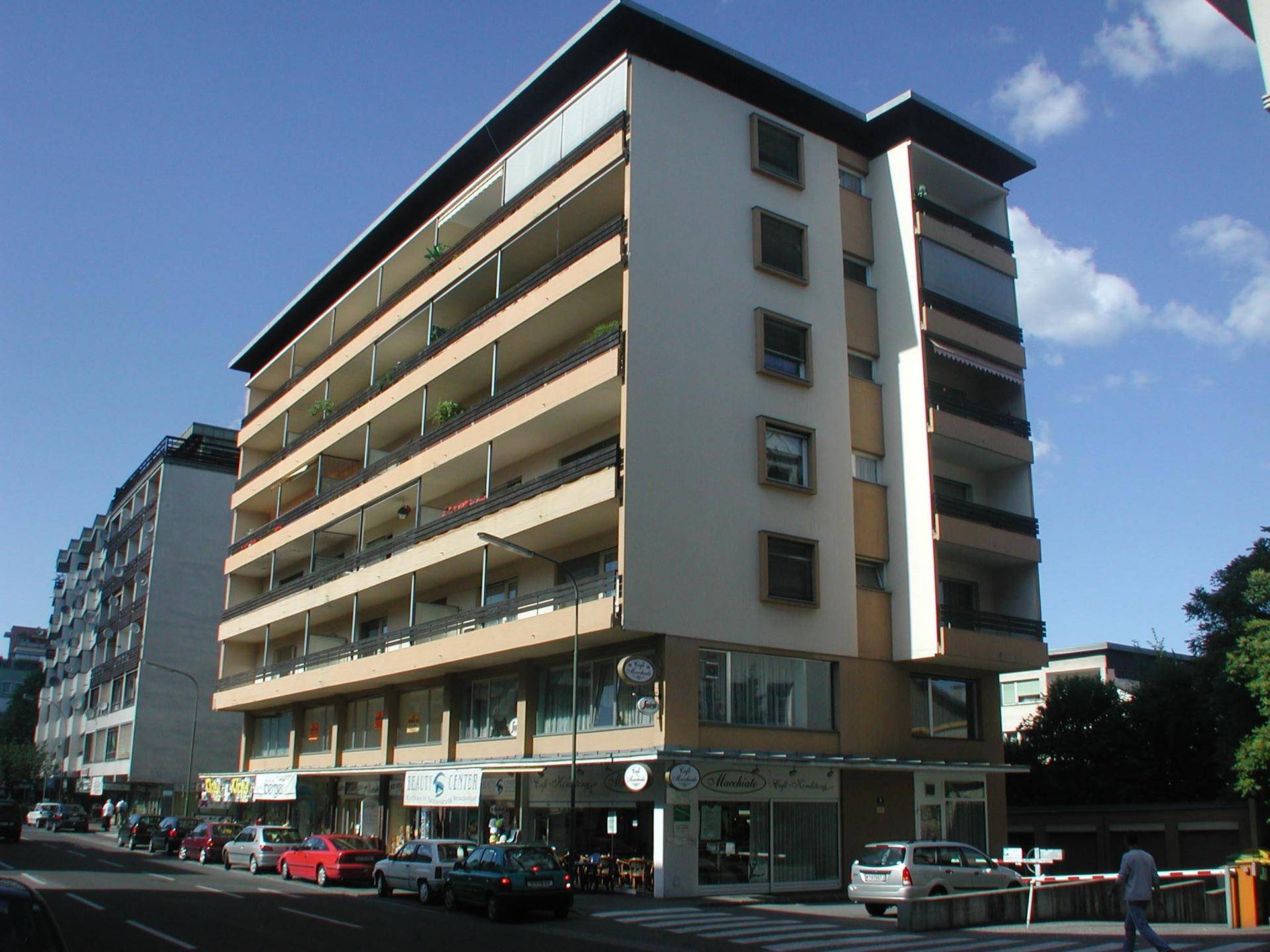 Mehrstöckiges Gebäude mit Balkonen und Geschäftsflächen im Erdgeschoss an einer belebten Stadtstraße.