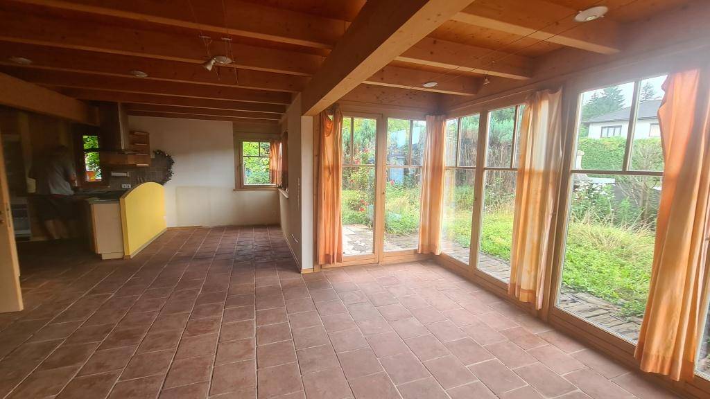 Geräumiger Wohnbereich mit Terrakotta-Fliesenboden, großen Fenstern und offener Küche im Hintergrund.
