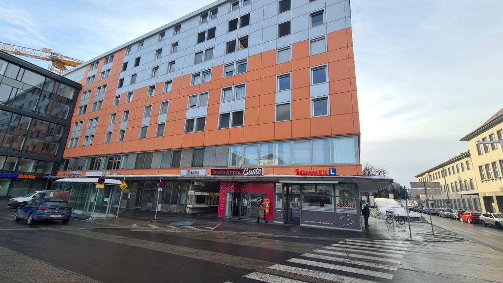 Modernes Geschäftsgebäude mit orangefarbener Fassade und Ladenlokalen im Erdgeschoss an einer Straße.