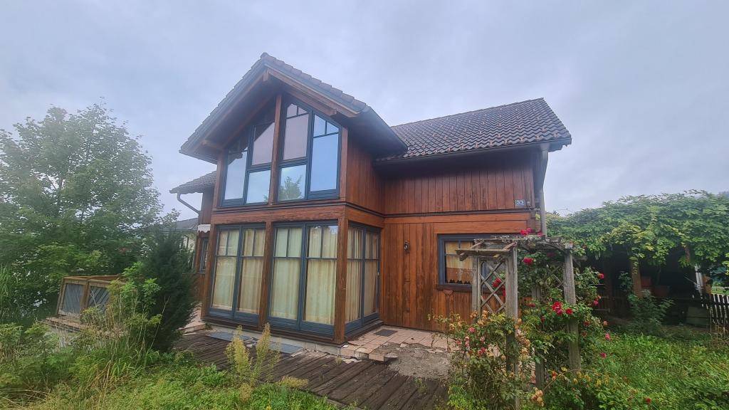 Charmantes Holzhaus mit großen Fenstern und traditionellem Satteldach, eingebettet in eine grüne Landschaft.