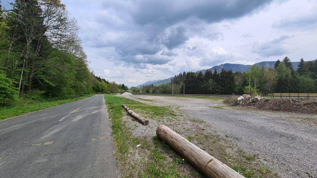 Unbebautes Grundstück entlang einer Landstraße mit Holzstämmen und bewaldeter Berglandschaft.