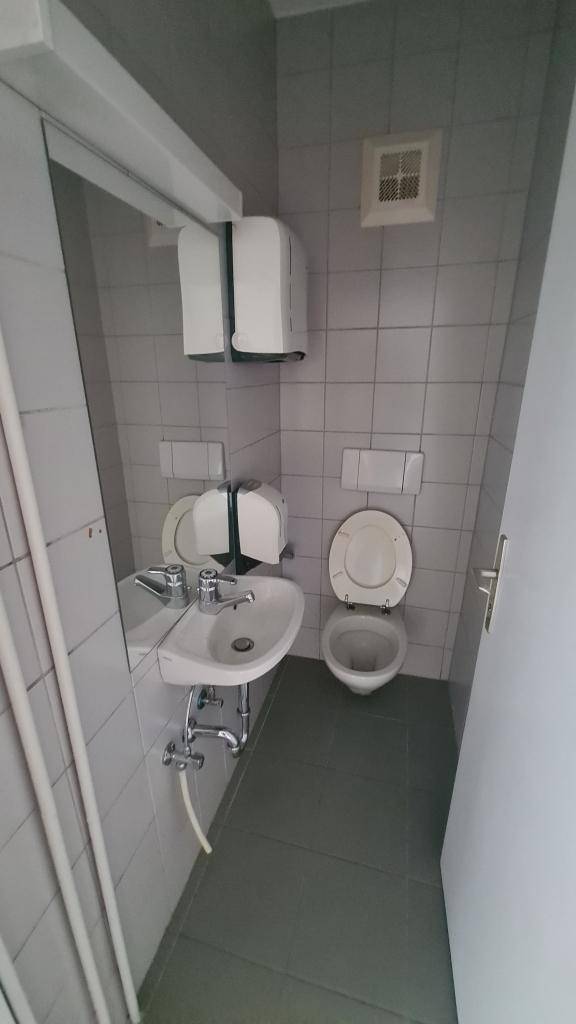 Kleiner Toilettenraum mit grauen Fliesen, WC, Waschbecken und Spiegel, ausgestattet mit einem Abluftventilator.