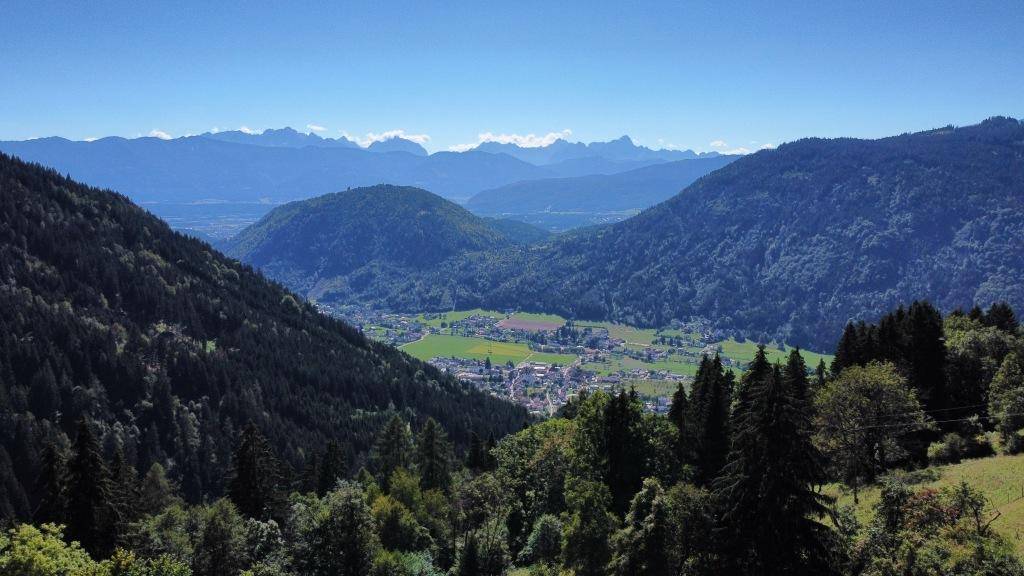 Panoramablick auf ein grünes Tal mit einer kleinen Stadt, umgeben von majestätischen Bergen und Wäldern.