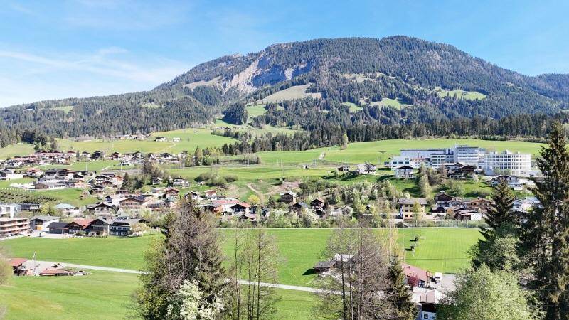 Weitläufiger Blick über eine ländliche Siedlung mit bewaldeten Bergen und grünen Wiesen.