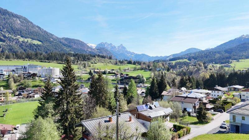 Panoramablick auf eine Wohngegend mit grünen Hügeln, schneebedeckten Gipfeln und Solaranlagen auf Dächern.