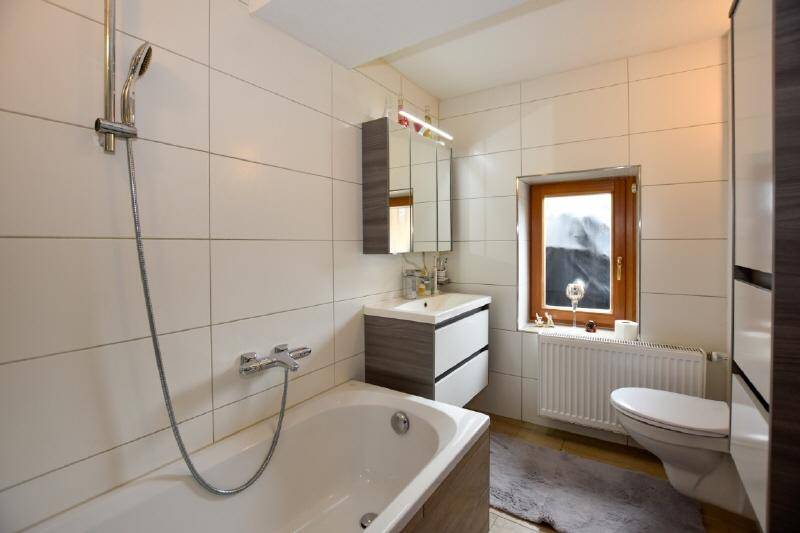 Helles Badezimmer mit weißen Fliesen, Badewanne mit Duschfunktion und einem Fenster. 