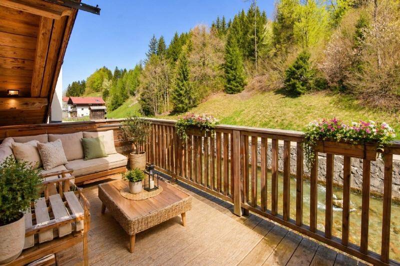 Gemütlicher Holzbalkon mit Sitzgelegenheiten und idyllischem Blick auf den Bach und die Berge.
