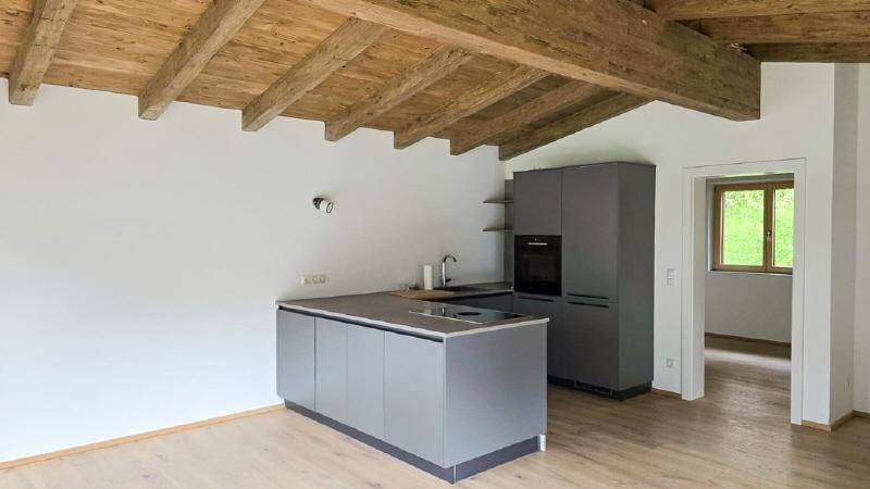 Offener Wohnbereich mit moderner grauer Küchenzeile, Holzbalkendecke und hellem Parkettboden.