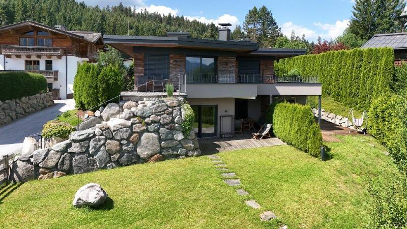 Modernes Haus mit Steinfassade, Balkon und gepflegtem Garten in alpiner Umgebung.