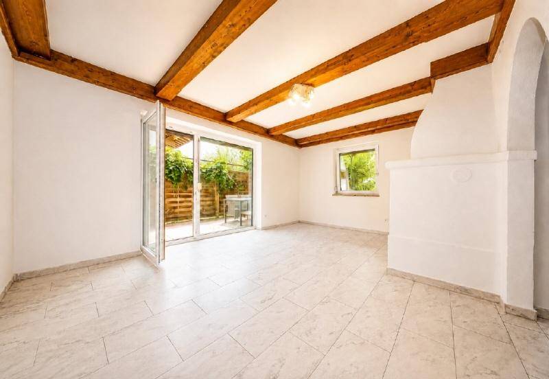 Heller Wohnraum mit sichtbaren Holzbalken, Fliesenboden und Zugang zu einer Außenterrasse.