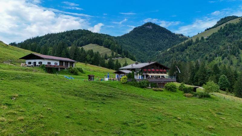 Zwei traditionelle Gebäude auf einem grünen Berghang, umgeben von Wäldern und hohen Bergen.