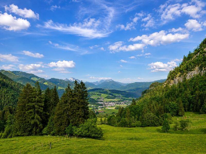 Weitläufige Landschaft mit grünen Hügeln, bewaldeten Bergen und einem Tal mit einem Dorf unter blauem Himmel.