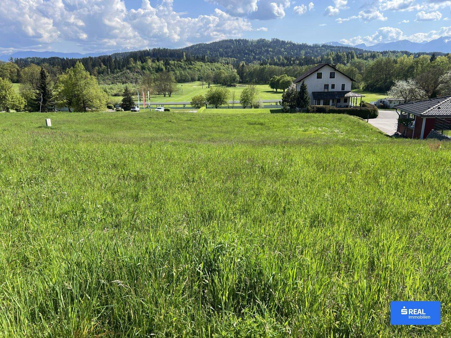 Sonnige Wiesenfläche mit Blick auf bewaldete Hügel und vereinzelte Häuser in der Ferne.