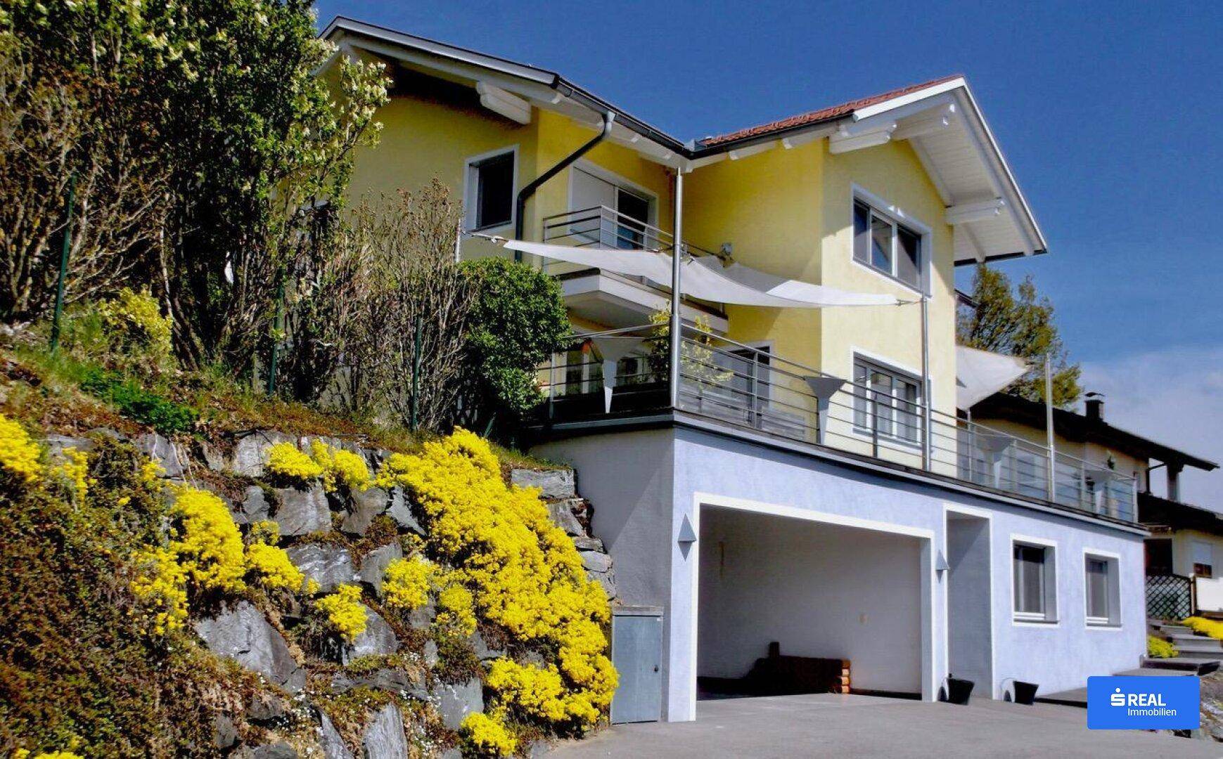 Modernes Haus mit gelber Fassade, mehreren Balkonen und integrierter Doppelgarage, umgeben von Grün.