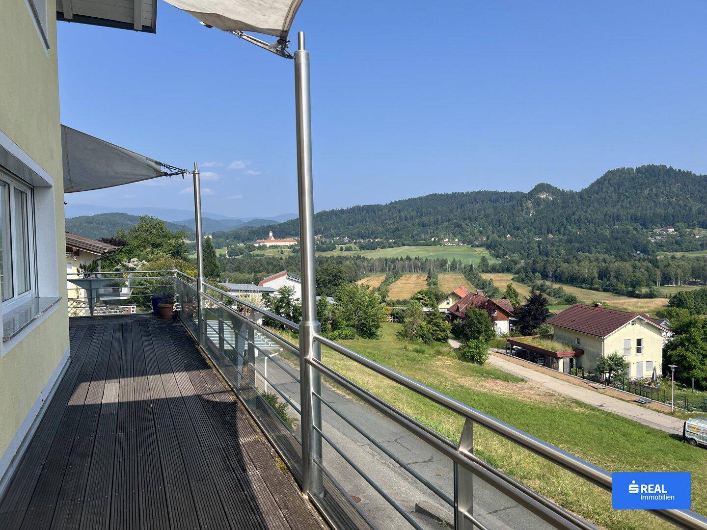 Breiter Balkon mit Glasgeländer und Panoramablick über die grüne Landschaft und ferne Berge.