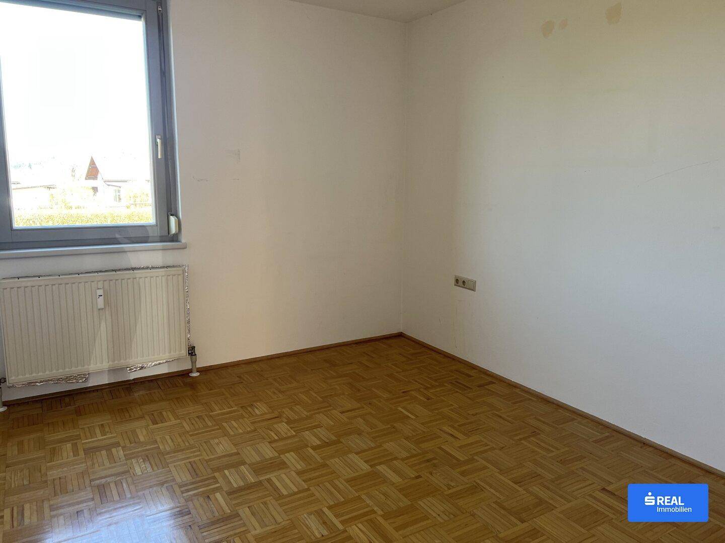 Helles Zimmer mit Parkettboden, Fenster und Heizkörper.