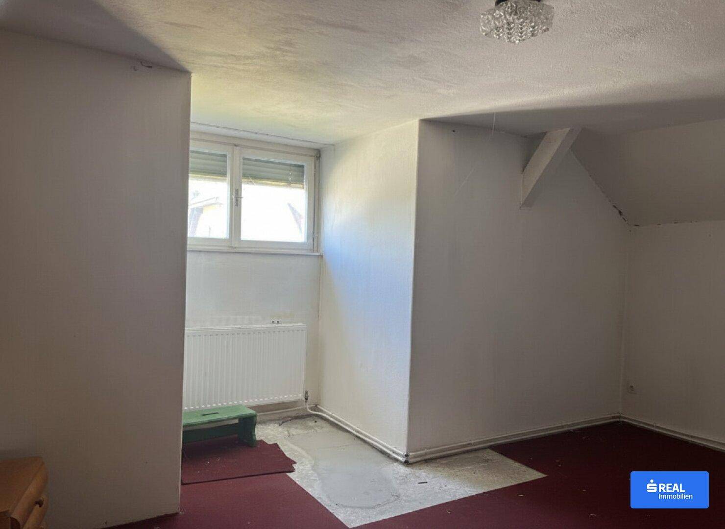 Leerer Raum mit Fenster und Heizkörper, der Renovierungsbedarf am Boden aufweist.