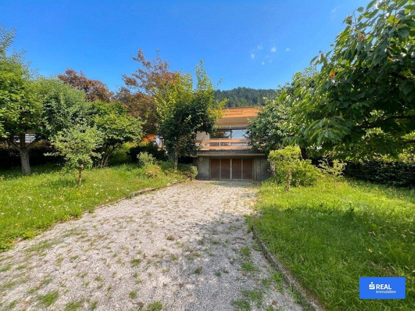 Modernes Haus mit Kiesauffahrt, umgeben von viel Grün und mit Bergblick unter blauem Himmel.