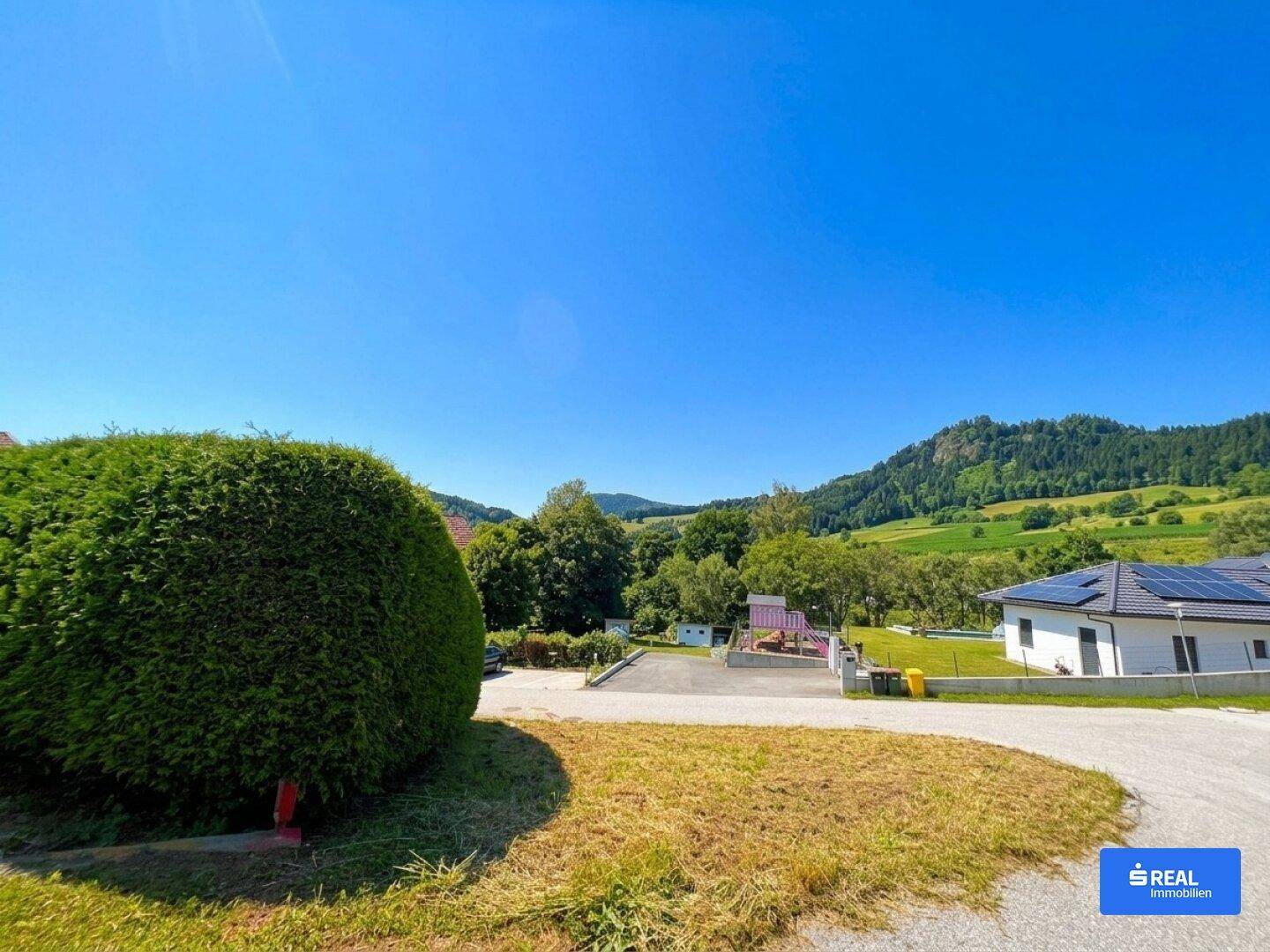 Wohngegend mit einem Spielplatz, gepflegter Hecke und einem Haus mit Solaranlage in ländlicher Umgebung.
