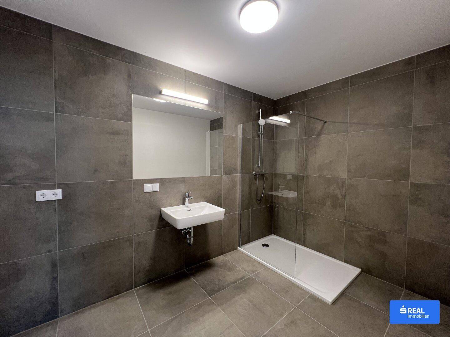 Modernes Badezimmer mit dunklen Fliesen, wandhängendem Waschbecken und ebenerdiger Dusche.
