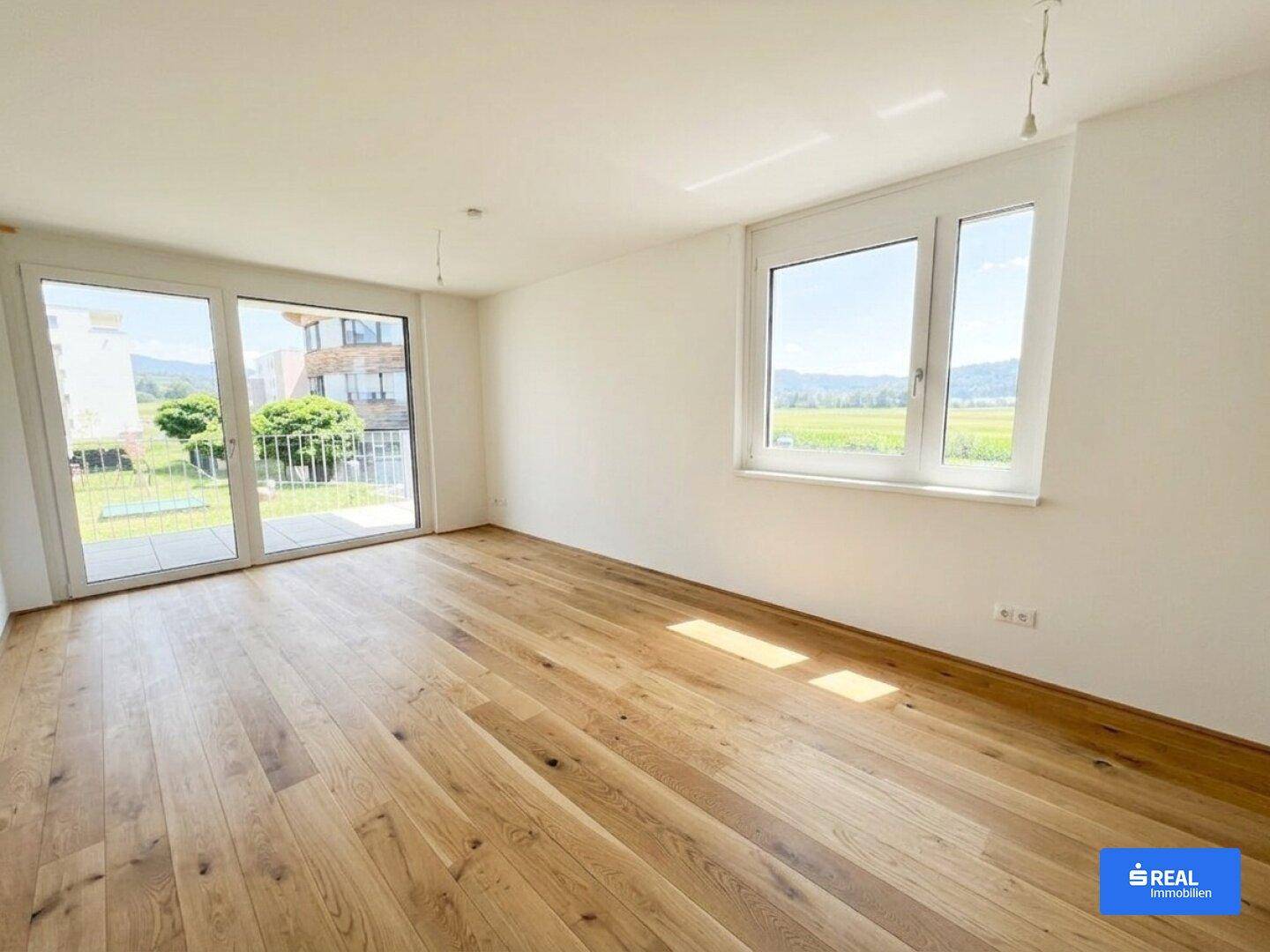 Heller Raum mit Holzboden, großen Fenstern und weitem Blick ins Grüne und auf Berge.