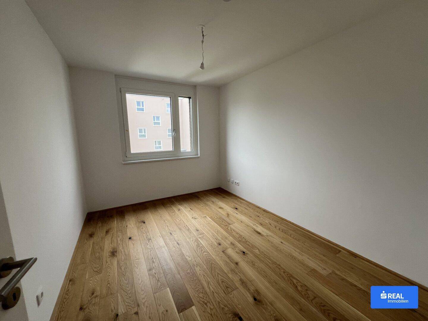 Helles Schlafzimmer mit Holzparkettboden und großem Fenster, bereit zur individuellen Gestaltung und Nutzung.
