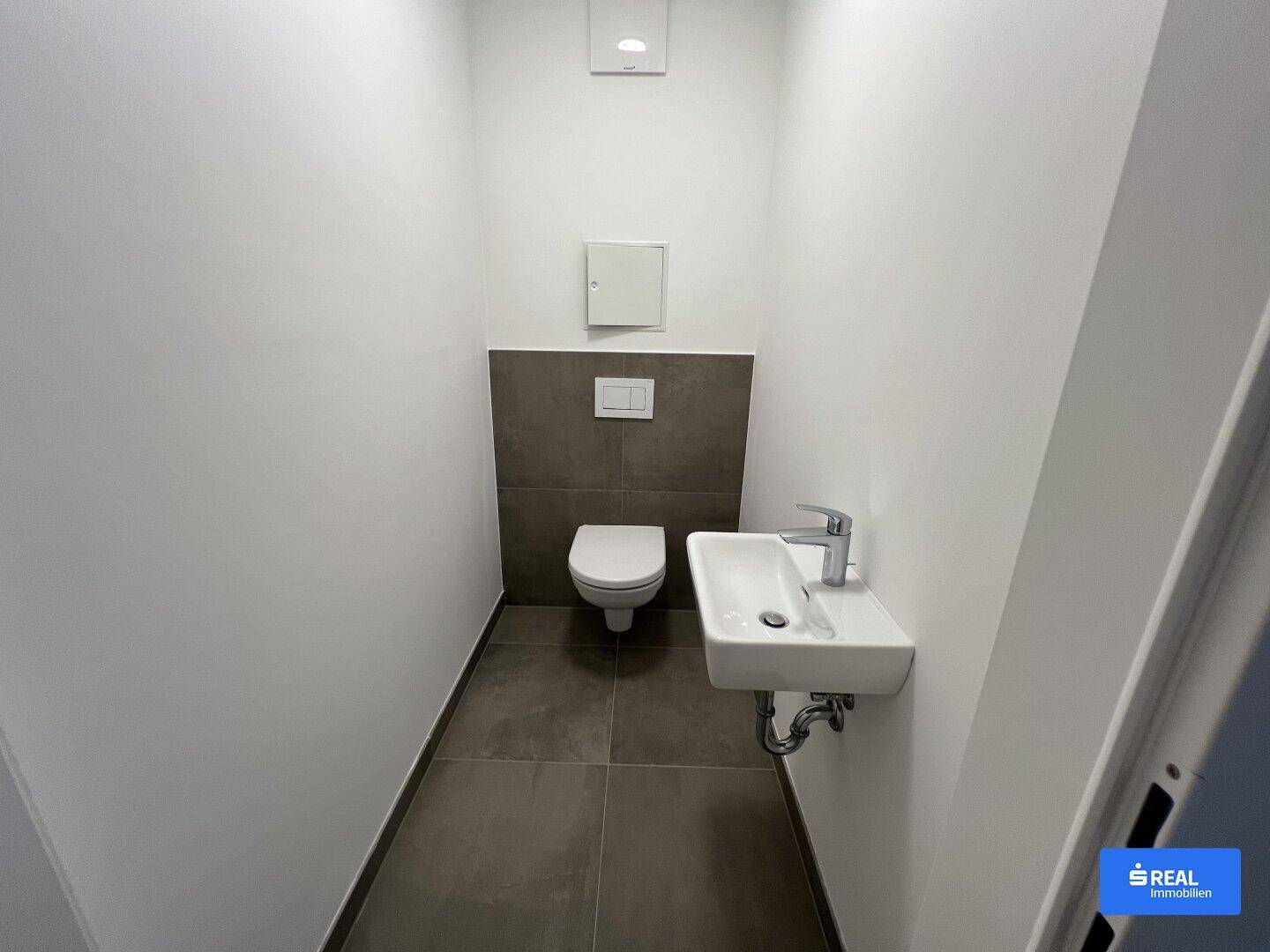 Modernes Gäste-WC mit Wand-WC, kleinem Waschbecken und dunklen Bodenfliesen in minimalistischem Design.