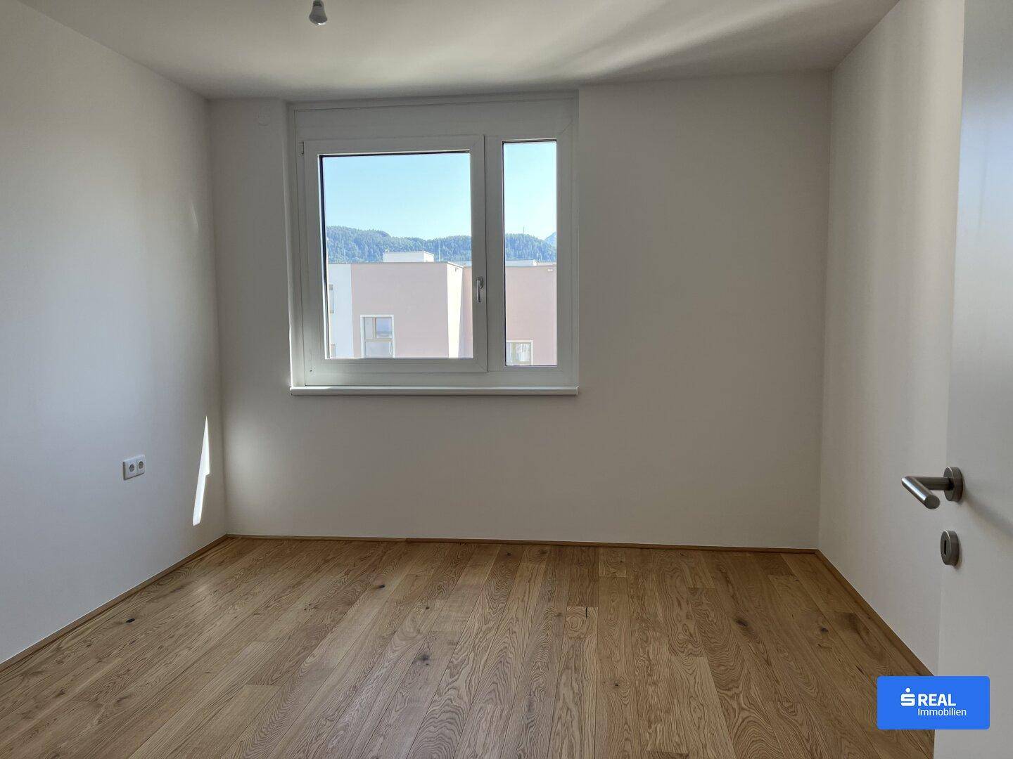 Leeres Schlafzimmer mit Holzboden und einem großen Fenster, das viel Tageslicht hereinlässt.