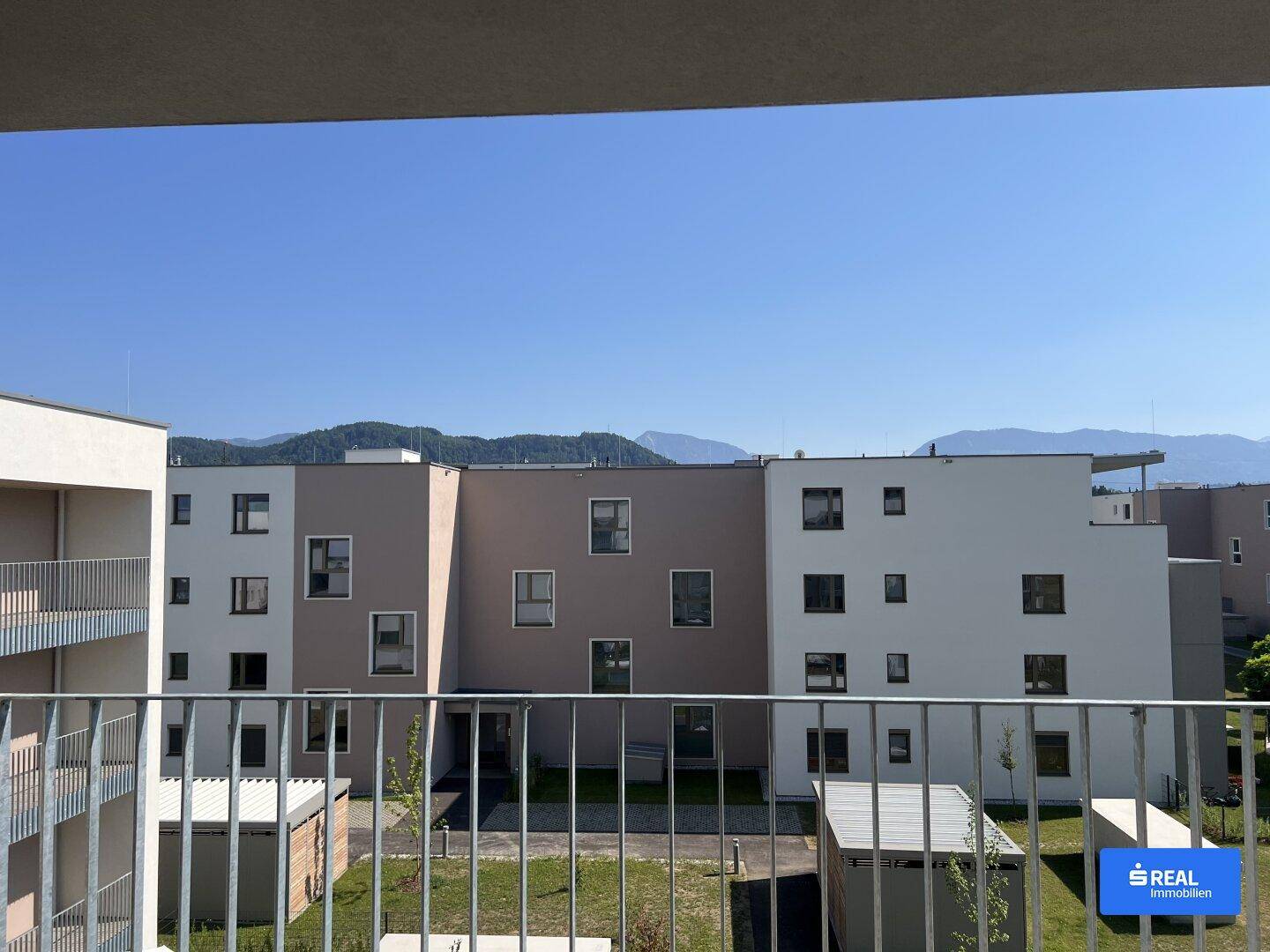 Blick von einem Balkon auf weitere moderne Gebäude und eine Berglandschaft im Hintergrund.