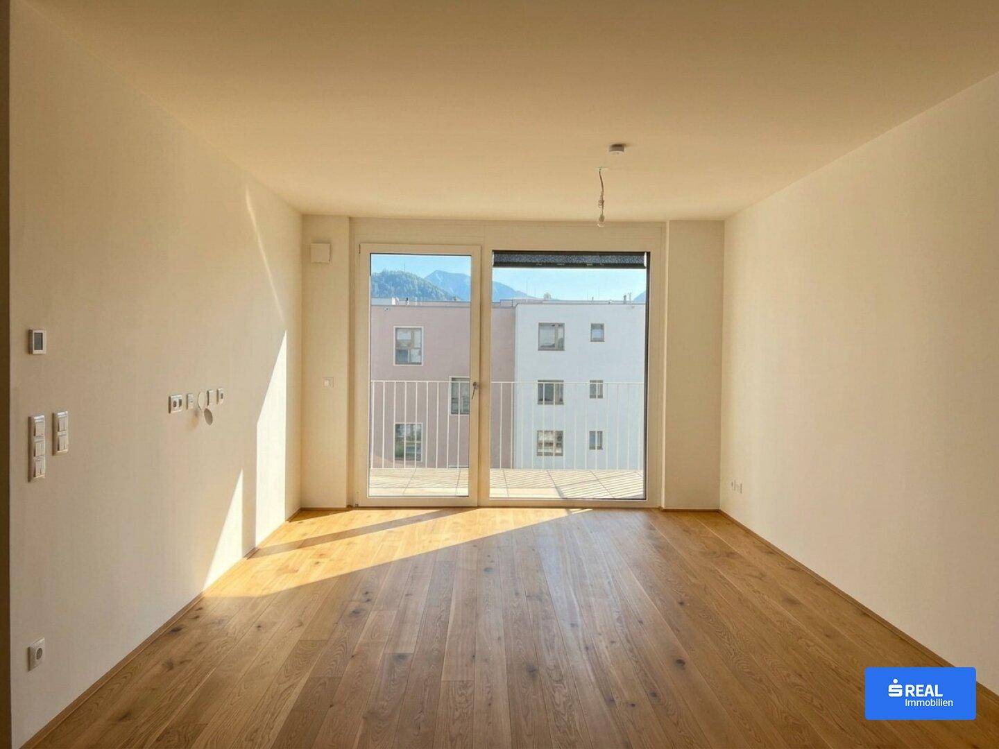 Helles, leeres Zimmer mit Holzboden und großem Fenster mit Zugang zu einem Balkon.