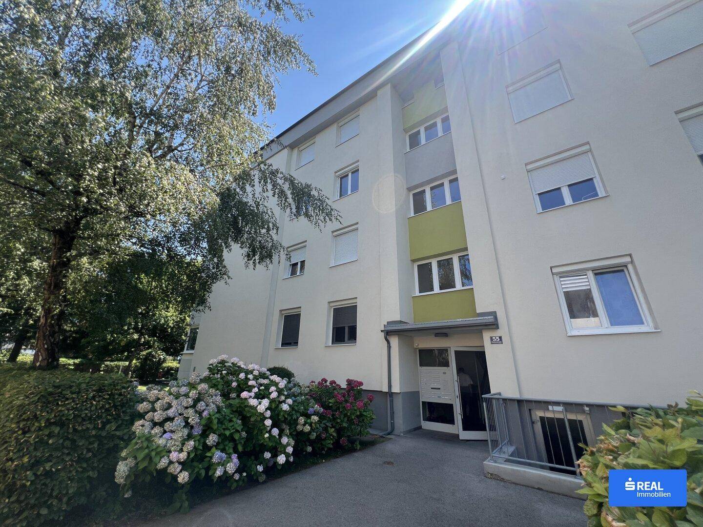 Eingangsbereich eines Mehrfamilienhauses mit blühenden Hortensien und viel Sonnenlicht.