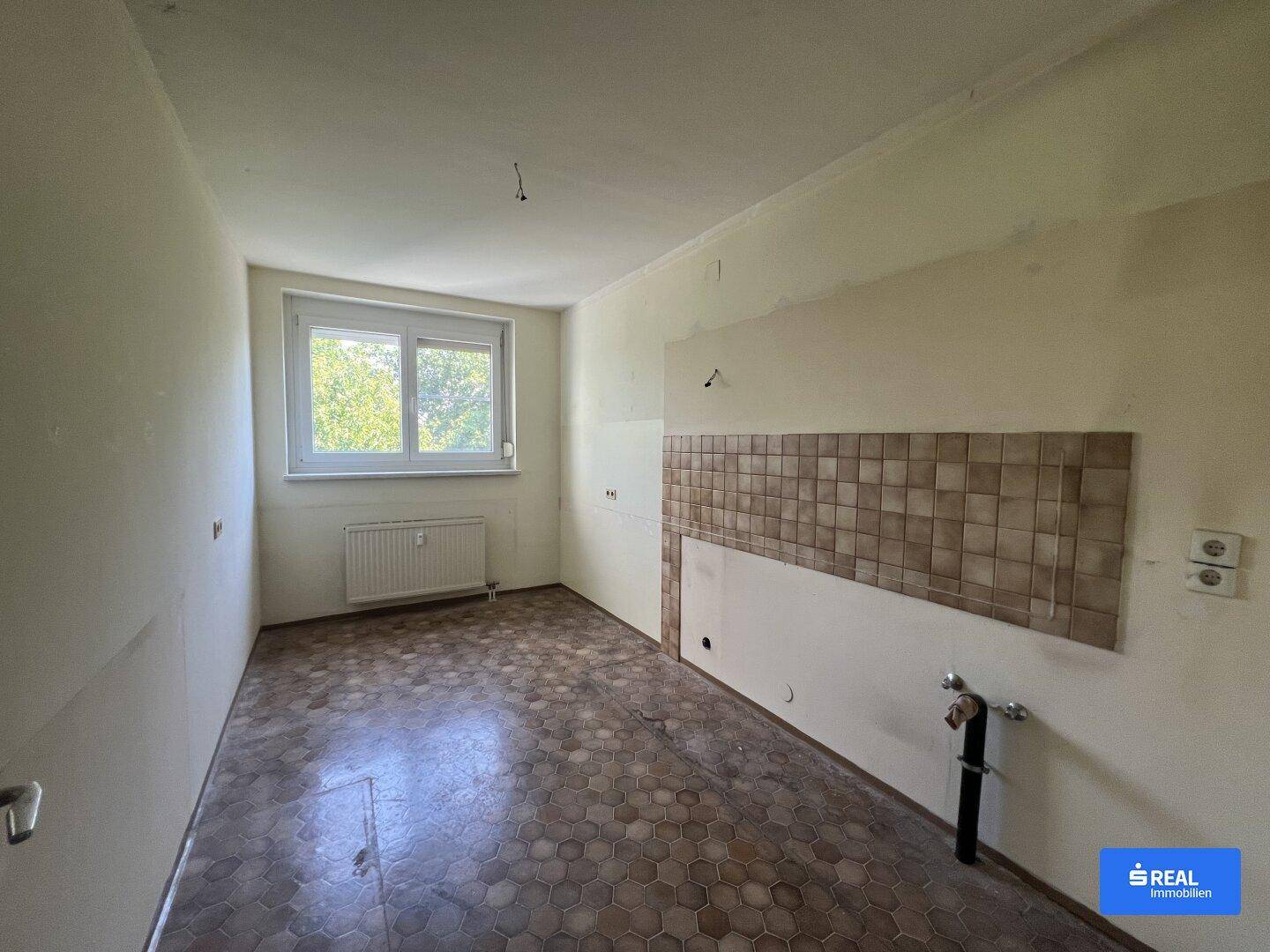 Leere Küche mit Fliesenboden, Fenster und vorbereiteten Anschlüssen für eine Einbauküche.