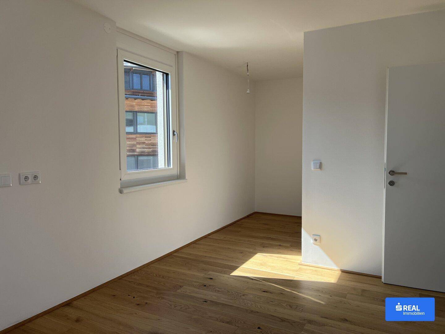 Sonniger Raum mit Holzboden und einem Fenster, das den Blick auf die umliegenden Gebäude freigibt.