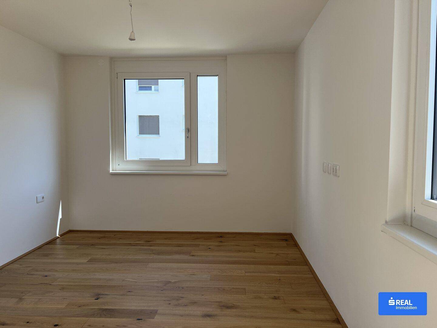 Geräumiges Zimmer mit Holzboden und zwei Fenstern, die für eine helle und freundliche Atmosphäre sorgen.