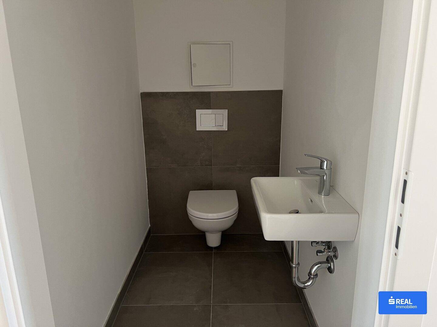 Kleines Gäste-WC mit Wand-WC, Handwaschbecken und dunklen Fliesen im unteren Wandbereich.
