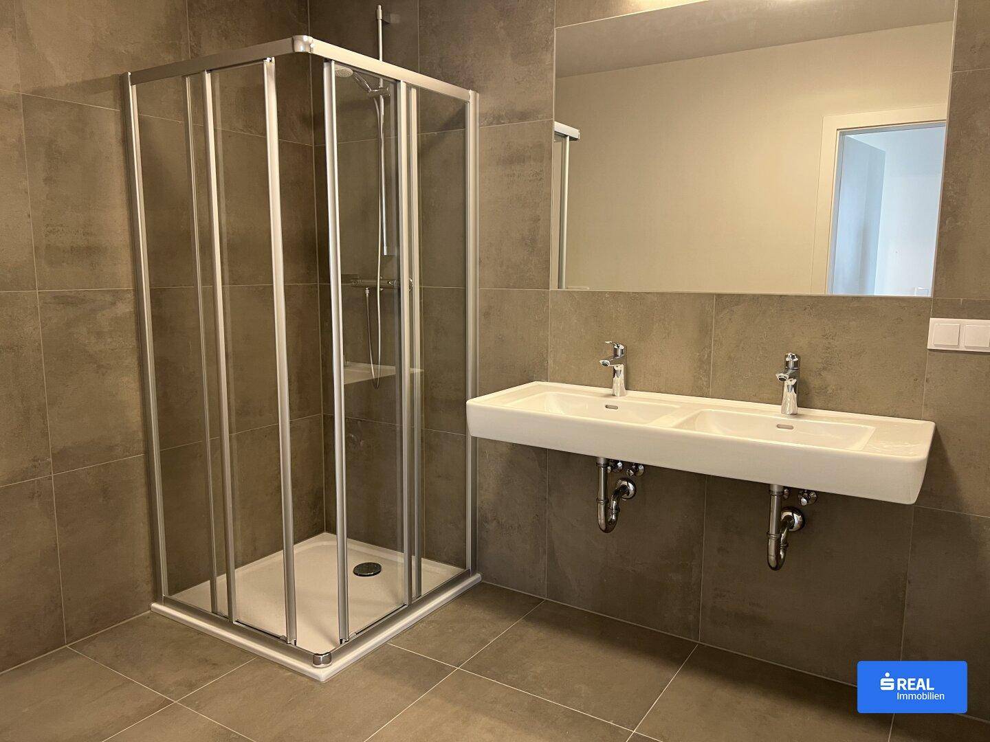 Modernes Badezimmer mit doppelten Waschbecken, großer Duschkabine und grauen Fliesen an Wänden und Boden.