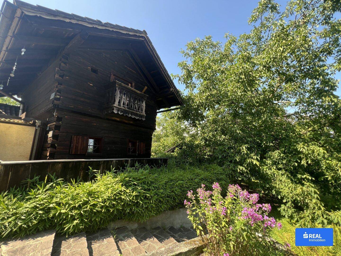 Traditionelles Holzhaus mit Balkon und üppiger Vegetation unter strahlend blauem Himmel.