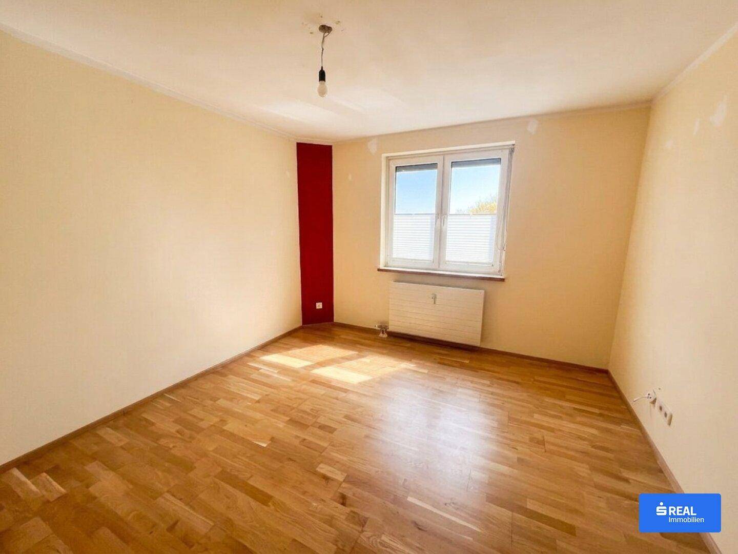 Leeres Schlafzimmer mit Parkettboden, einem Fenster und einer roten Akzentwand, bereit zur Gestaltung.