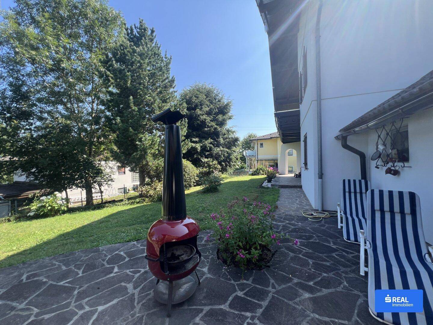 Sonniger Terrassenbereich mit Steinplatten, Liegestühlen und einem Grill im Garten.