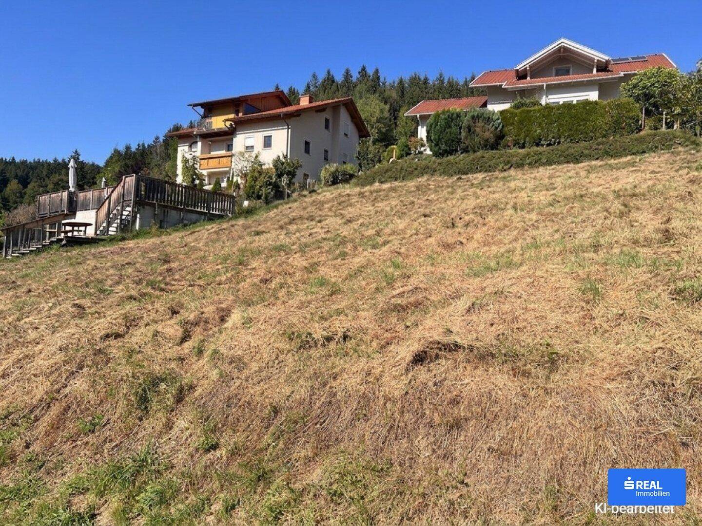 Sonniges, hügeliges Grundstück mit modernen Wohnhäusern und einer Holzterrasse im Hintergrund.