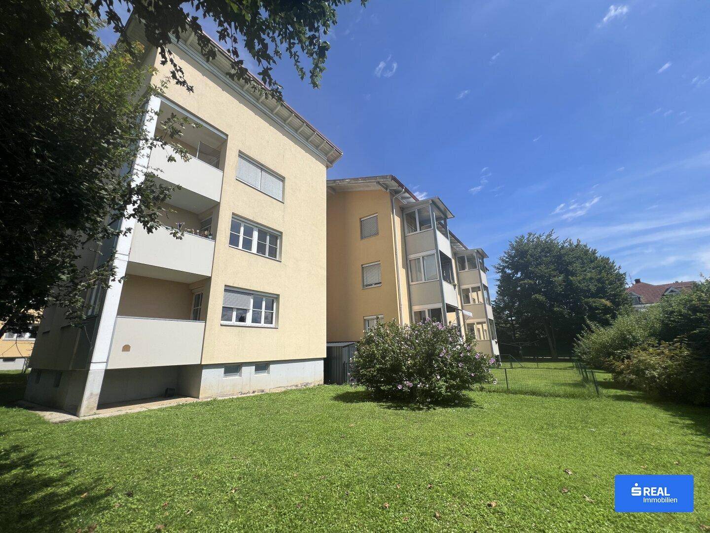 Gepflegte Grünfläche vor einem gelben Mehrfamilienhaus mit Balkonen unter blauem Himmel.