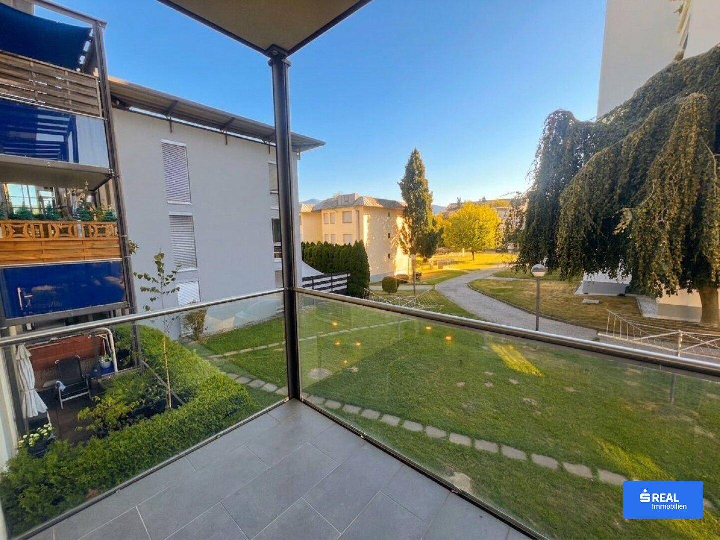 Balkon mit Glasgeländer und Blick auf die grüne Umgebung und einen Spazierweg unter blauem Himmel.