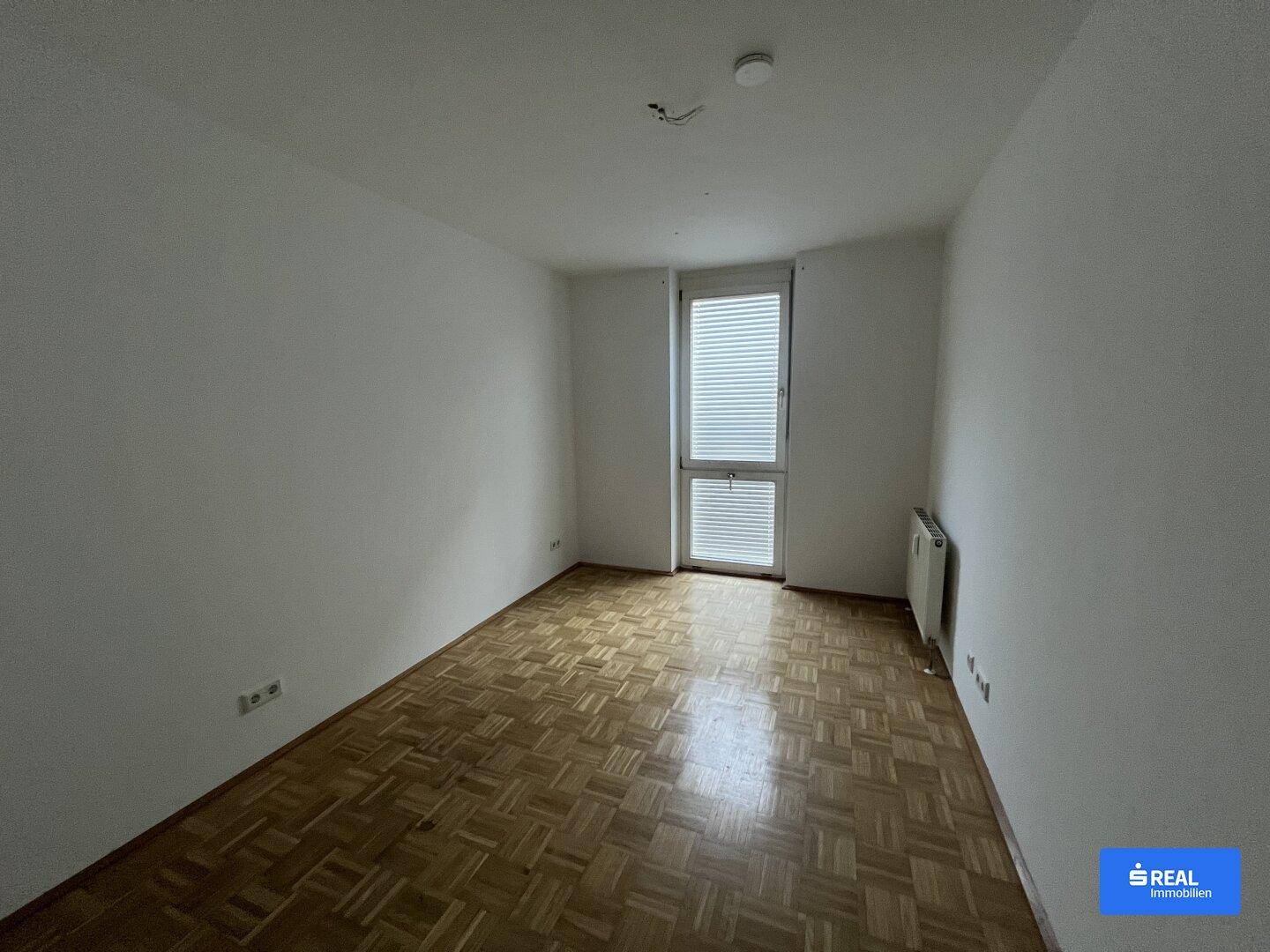 Helles Schlafzimmer mit Parkettboden und großem Fenster, das viel Tageslicht hereinlässt.