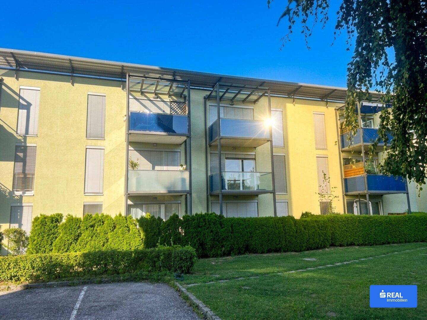 Nahaufnahme der modernen Fassade eines Mehrfamilienhauses mit Balkonen und gepflegten Hecken im Sonnenlicht.