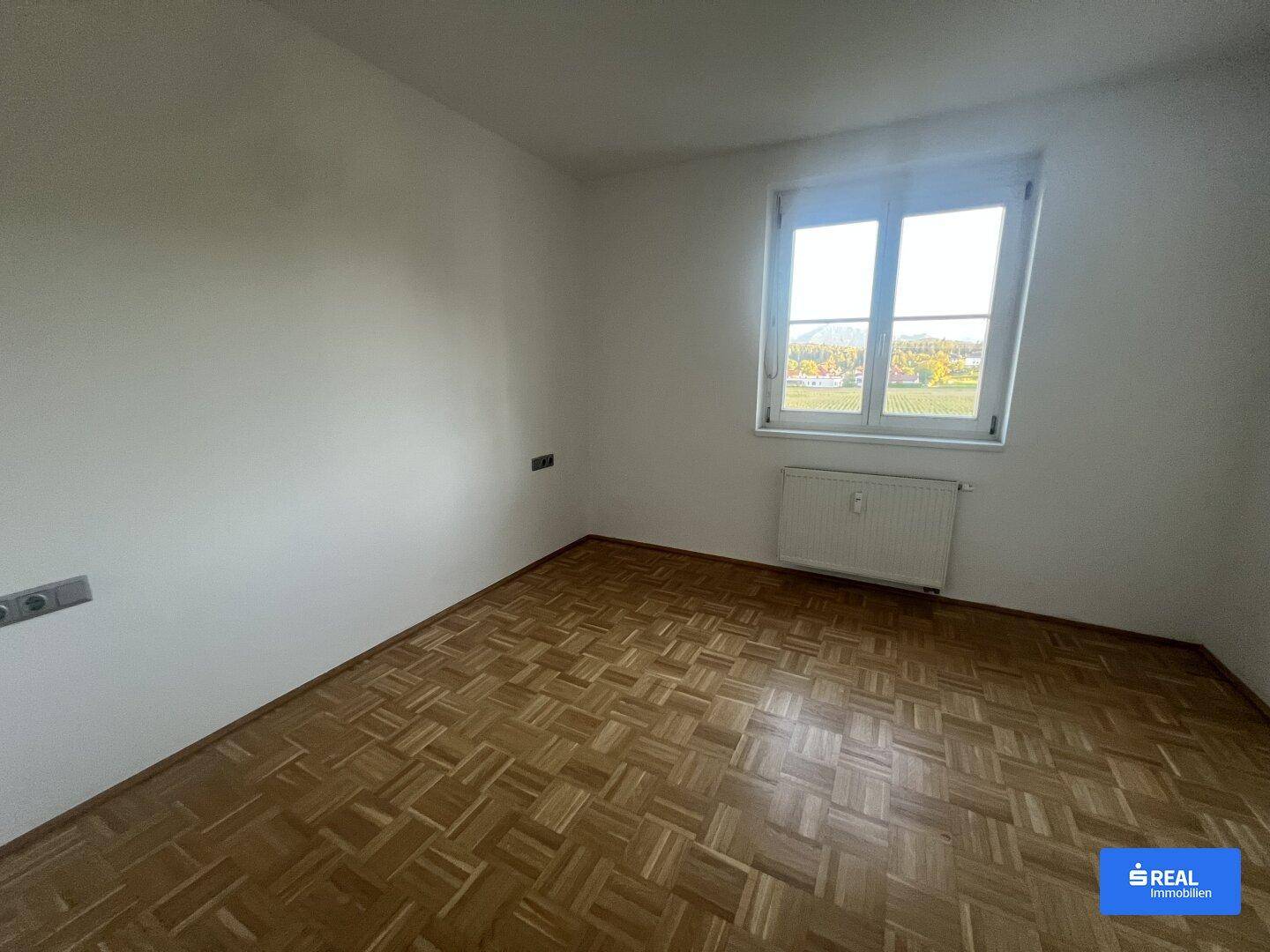 Helles Schlafzimmer mit Parkettboden und großem Fenster mit Blick ins Grüne.