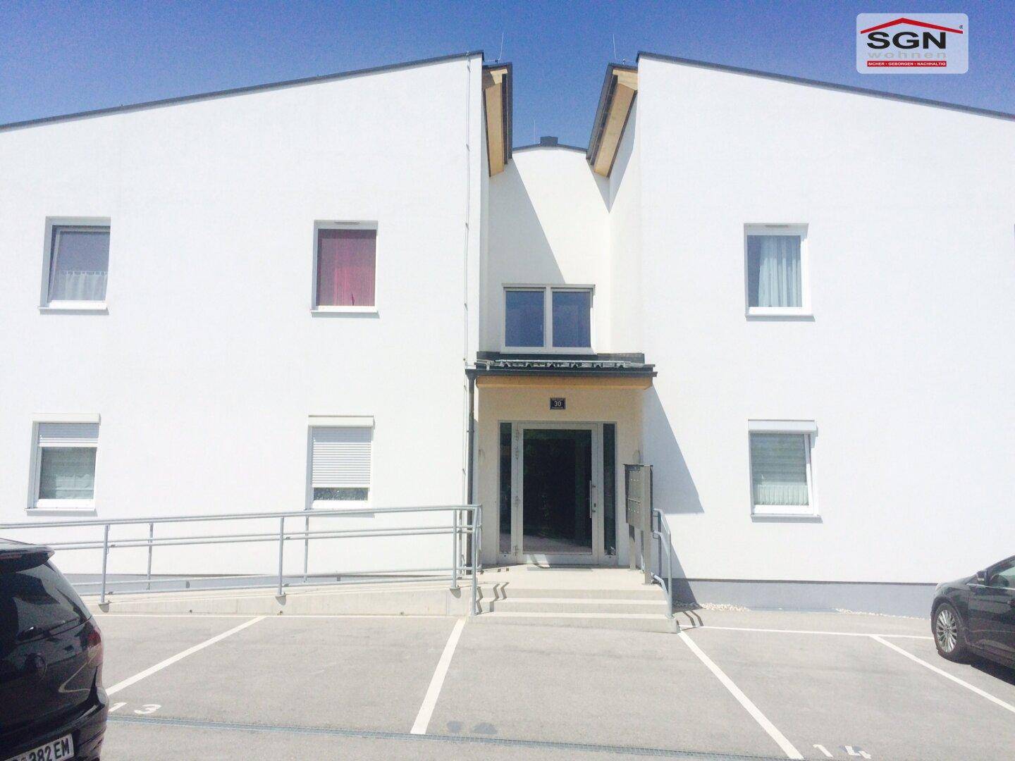 Eingangsbereich eines modernen Gebäudes mit Rampe und Parkplätzen davor.